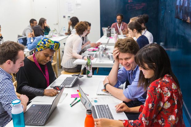 tables in a classroom, with people sitting around them looking at their laptops together