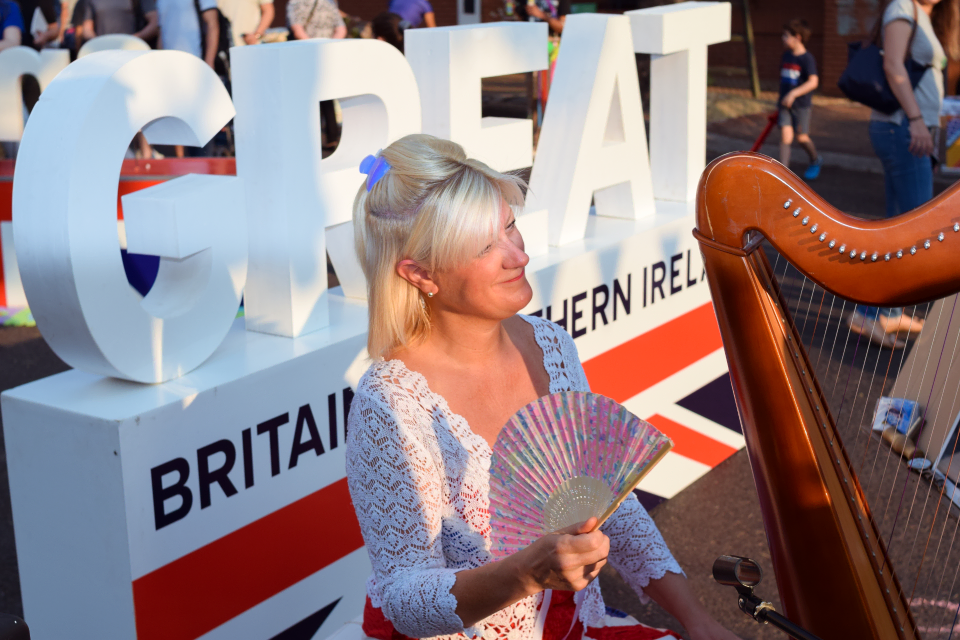 Harpist Anita Aslin fans herself next to her harp, in front of a GREAT Britain and Northern Ireland sign.