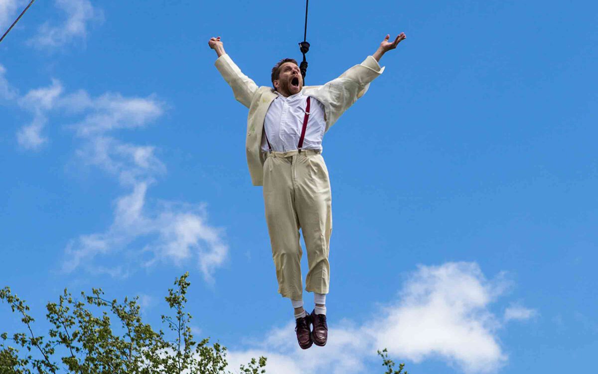 A male actor in a white suit and red braces is suspended in the blue sky, his arms outstretched and his mouth wide with glee.