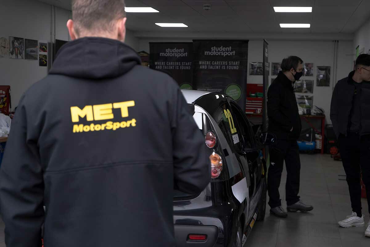 A MET student is stood in front a race car in a garage. The back of his jacket reads "MET Motorsport".