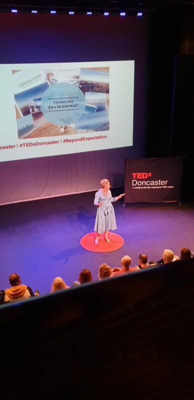 Lou Mycroft in a blue dress standing on the famous TED Talk red spotlight (actually a bathmat from Dunelm) at TEdxDoncaster 2019. Behind her is a slide representing the Thinking Environment.