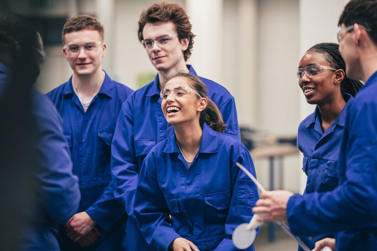 A young Indian woman, who is wearing protective eyewear, smiles as she enjoys her engineering class with her peers - they are all wearing blue coveralls.