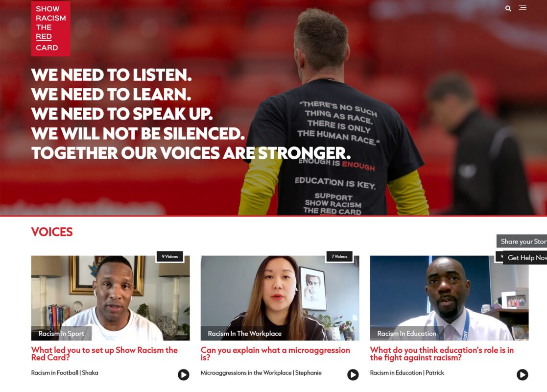 A footballer facing away from the camera towards the stands in a football stadium wearing a black top with the words in white that say - "There is no such thing as race There is only the human race" and pictures of two men and a woman underneath talking to camera about anti-racism