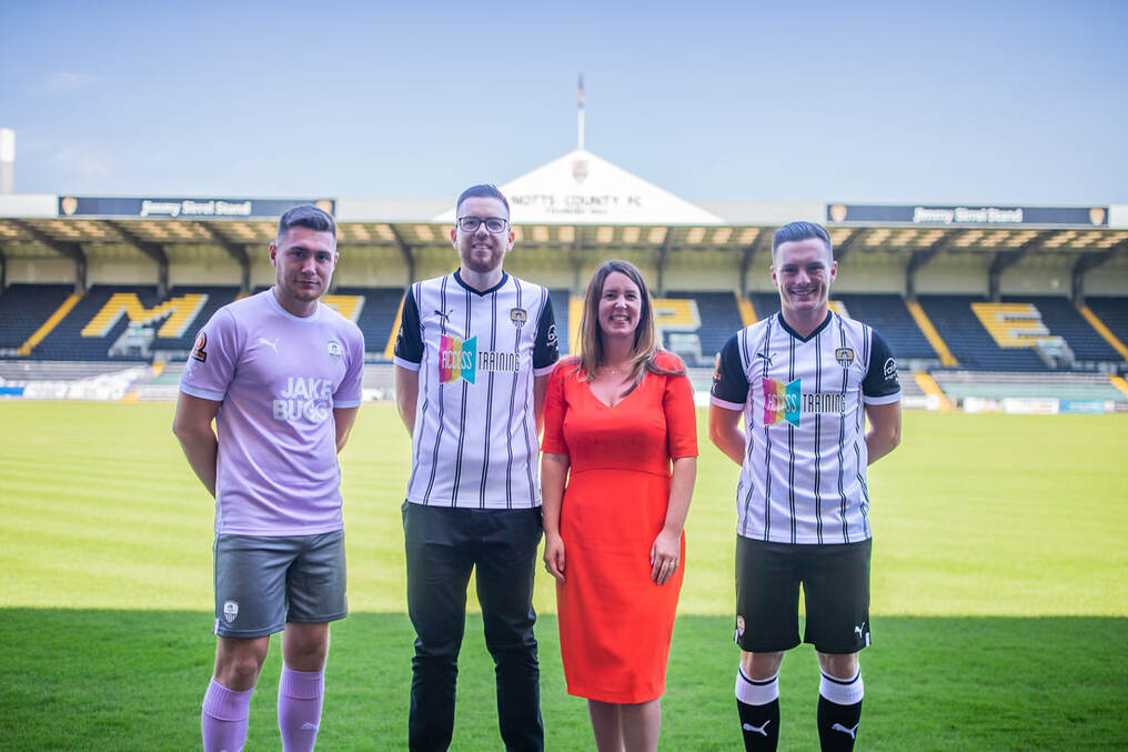 Four people standing on a football pitch wearing Notts County FC shirts