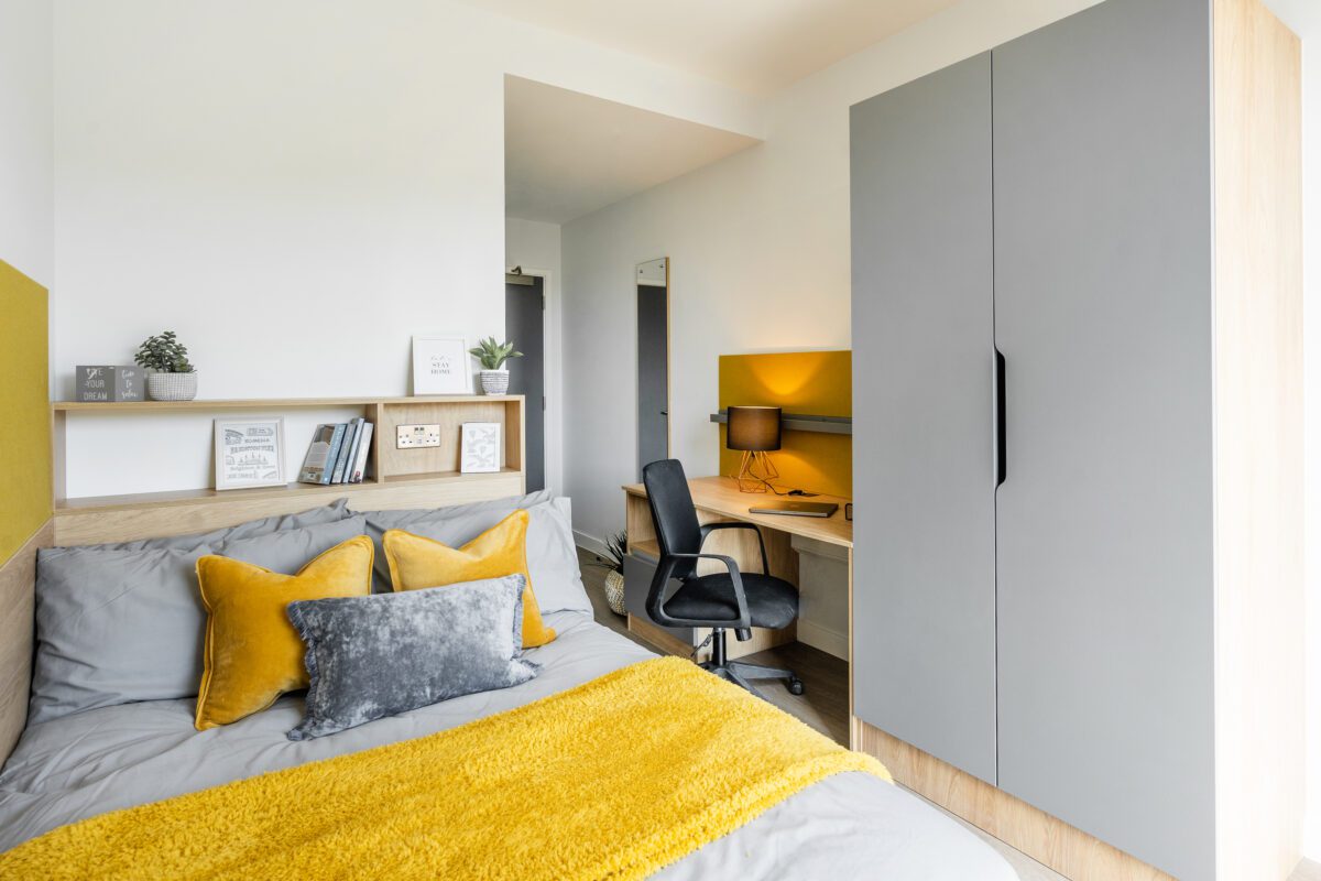 A cluster flat bedroom with a bed, wardrobe and desk. There is yellow and grey bedding and cushions, which coordinate with the wardrobe and desk pinboard.