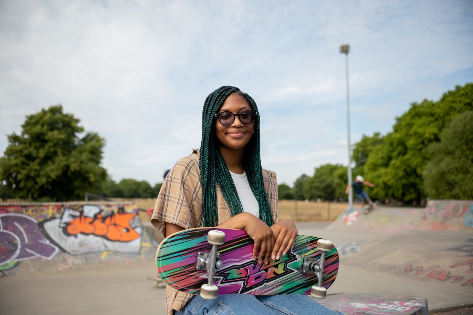 Image shows young woman in sunglases smiling at teh camera holding her skateboard