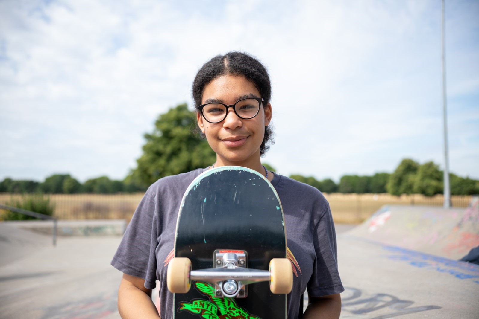Image shows young girl in glasses holding her skateboard looking at the camera