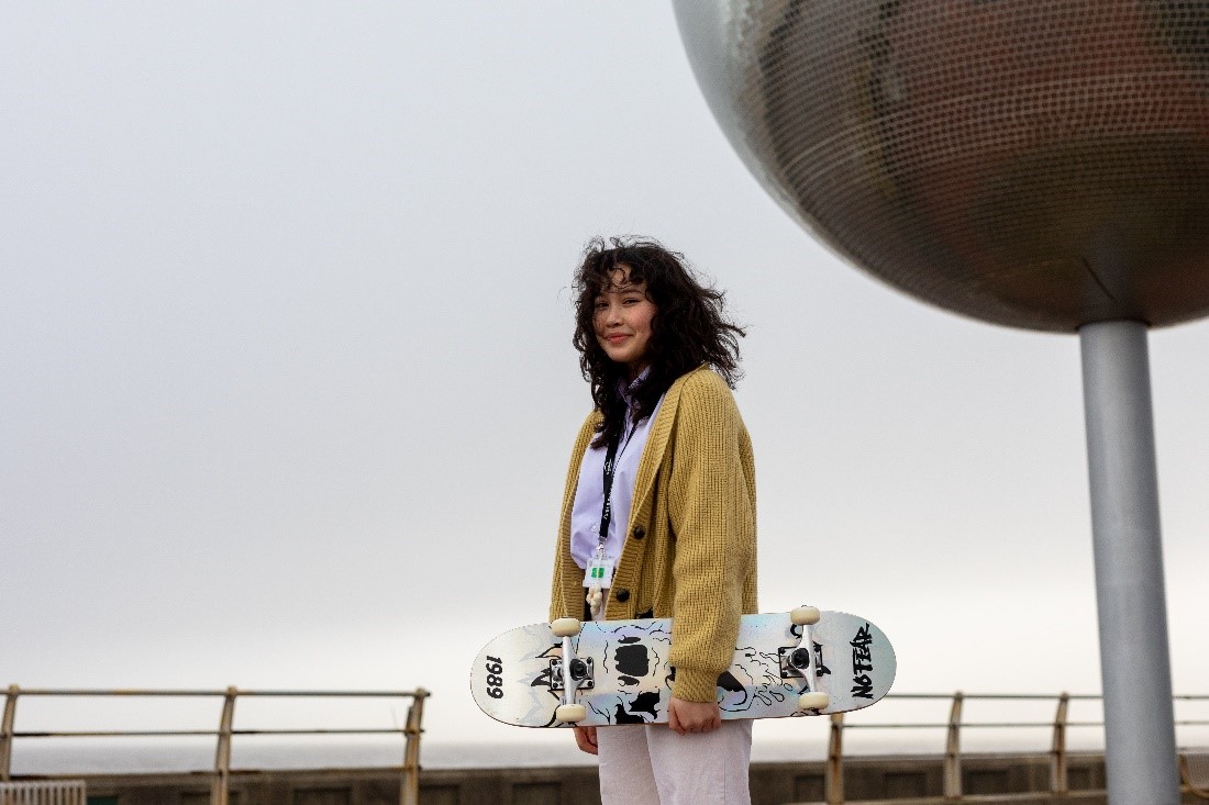 Image shows yougn woman smiling at camer holding her skatebaord with cloudy background