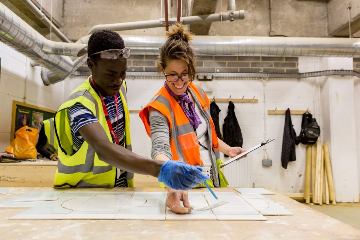 A teacher guides her student as they arrange tiles on a table in a construction workshop.
