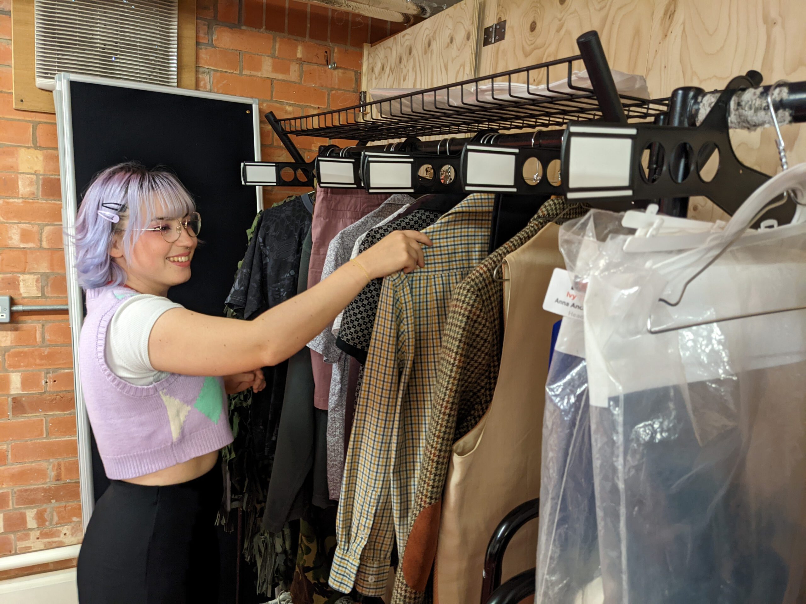 Young woman with purple hair hangs up a costume on a rail backstage at the theatre.