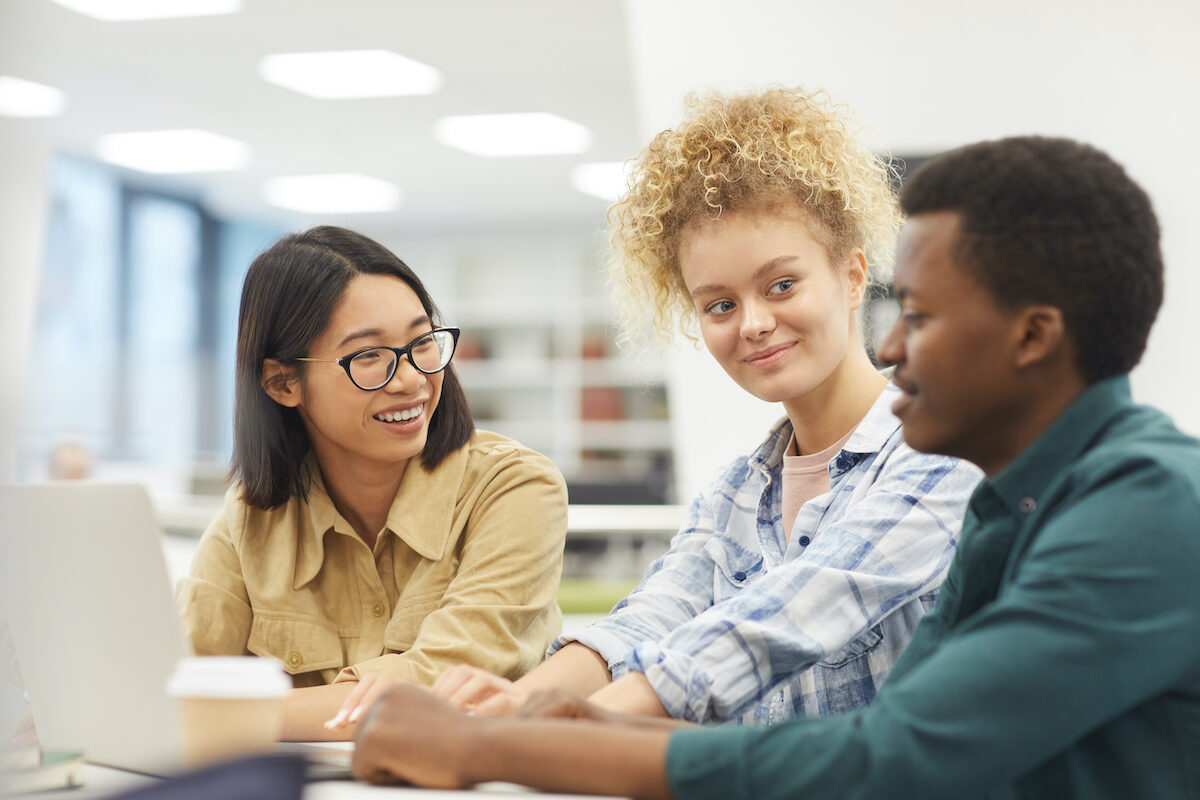 Multi-ethnic group of students using laptop together while discussing project in college library