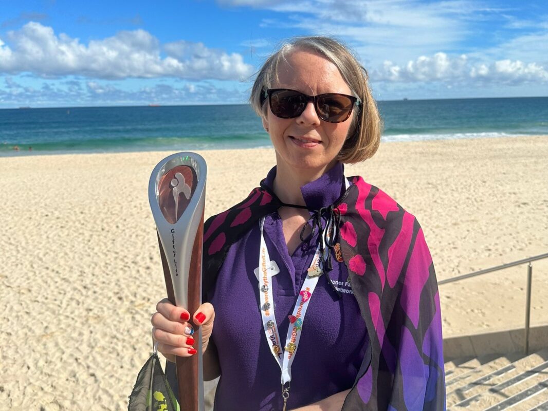 Bradford College staff member, Karen Piotr, holding a World Transplant Games 2023 baton while standing on the beach in Australia.