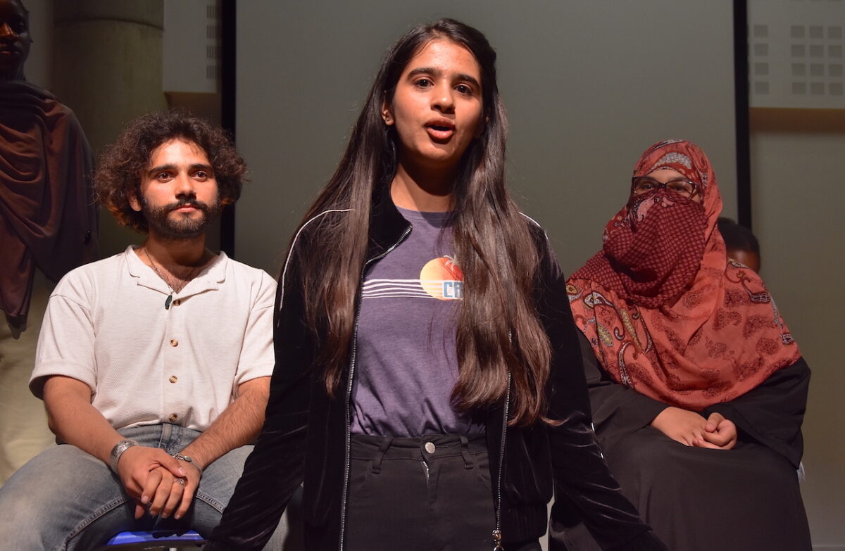 A Bradford College ESOL student speaks to the audience during a performance of ‘Because’ in Refugee Week 2023 while two other ESOL students watch from behind.