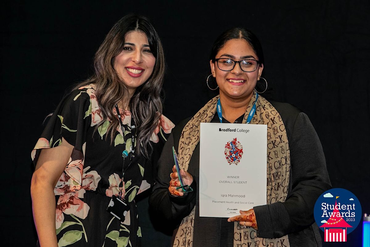 Alina Khan, Vice Principle for Student Experience, stands to the left of Iqra Mahmood, a Bradford College Health and Social Care student, as she collects her glass trophy and certificate on stage.
