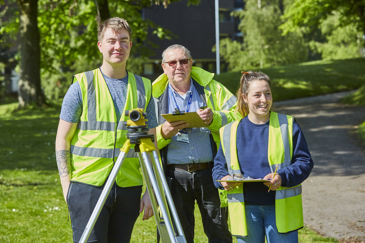 Two Leeds College of Building apprentices in high-viz jackets stand outside with their tutor using surveying equipment.