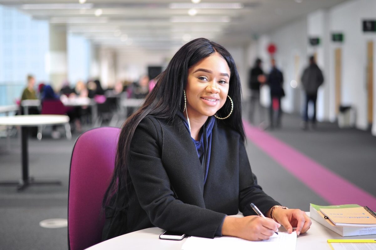 A female Bradford College student sits at a desk in a library and smiles at the camera.