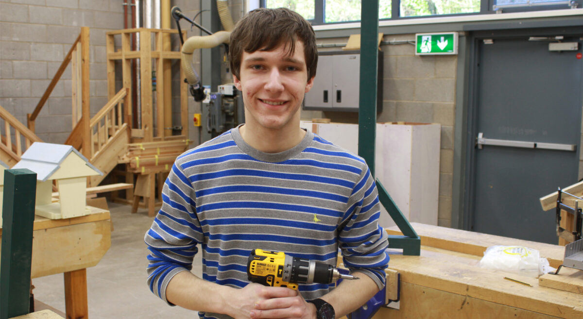 Young male smiling to camera holding a drill in a carpentry workshop.
