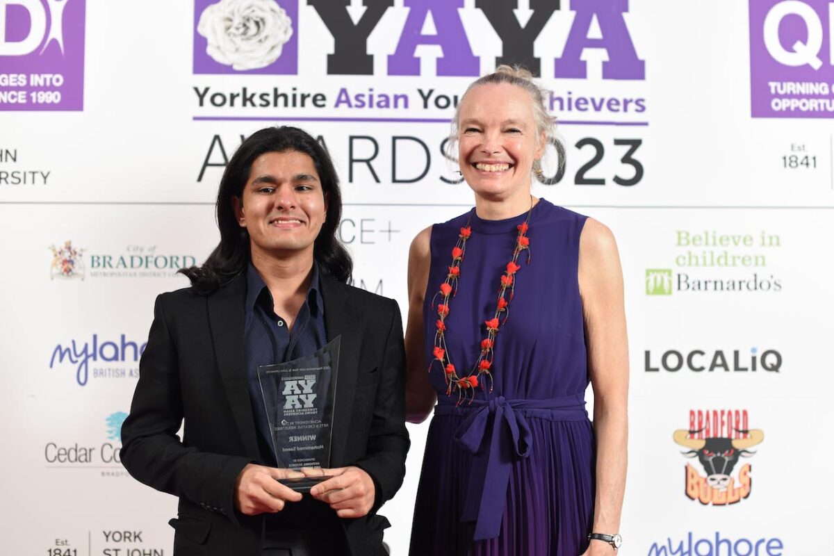 Student Mohammed Saeed stands to the left of Jo Quinton Tulloch, Director of the National Science and Media Museum. They both stand in front of a YAYA backdrop and he holds his glass trophy proudly.