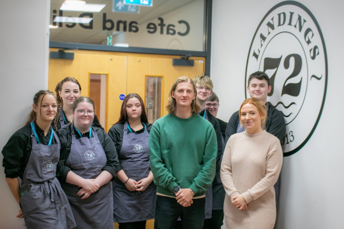 Cafe Culture students pose for a photo with representatives from coffee brothers, stood in front of double doors and a white wall with a decal that reads Landings 72