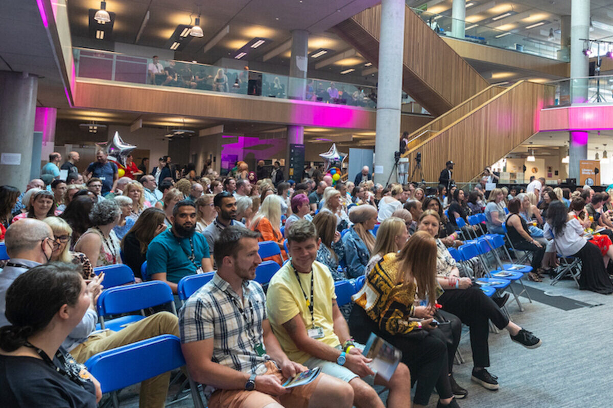 Hundreds of Bradford College staff are seated in a large campus building atrium awaiting a presentation at the staff conference.