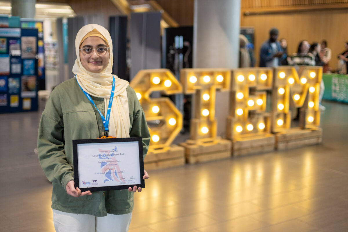 Bradford College biology student, Laibba Arshad Mahmood Sadiq, stands in front of a lit STEM sign holding her bronze British Biology Olympiad certificate.