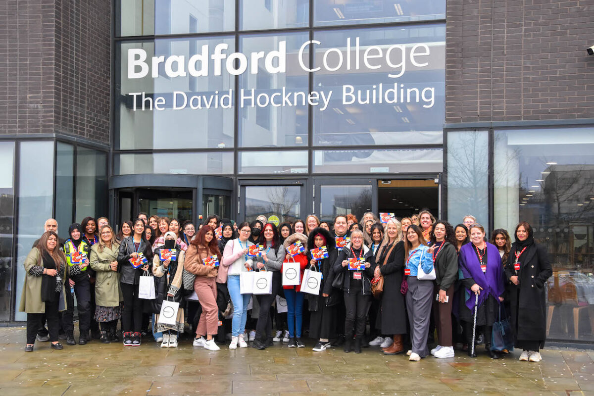 A large group of female staff and students pose outside the entrance of the Bradford College David Hockney Building holding Women of the World (WOW) Festival merchandise.