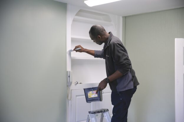 A male Leeds College of Building students stands on a ladder while painting a recessed bookshelf at Herd Farm.