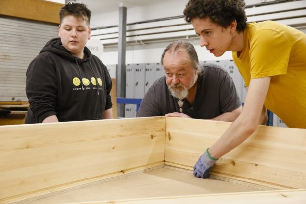 Two students stand either side of their tutor in the Leeds College of Building carpentry workshop and hold pieces of wood being joined together.