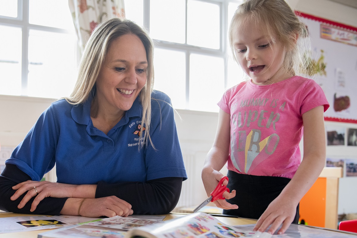 Nursery worker smiling while a young child holding scissors cuts out a magazine