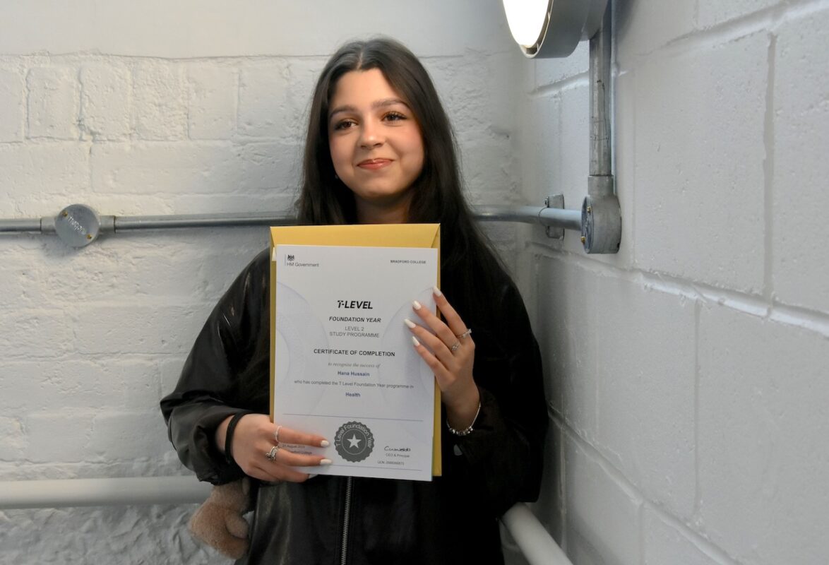 A Bradford College student, Hana Hussain, stands in front of a white wall proudly holding her T Level results certificate and envelope.