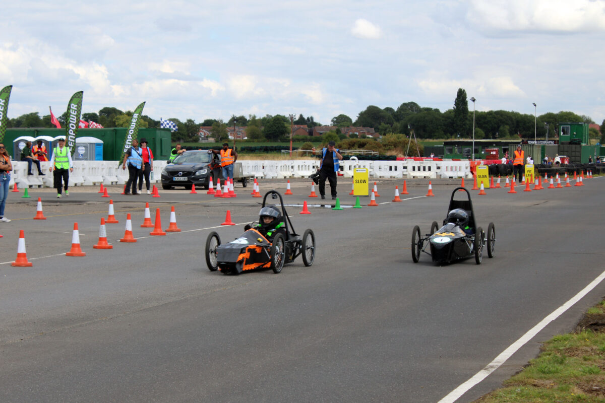 Two electric kit cars driven by school pupils in helmets drive around an outdoor race track.