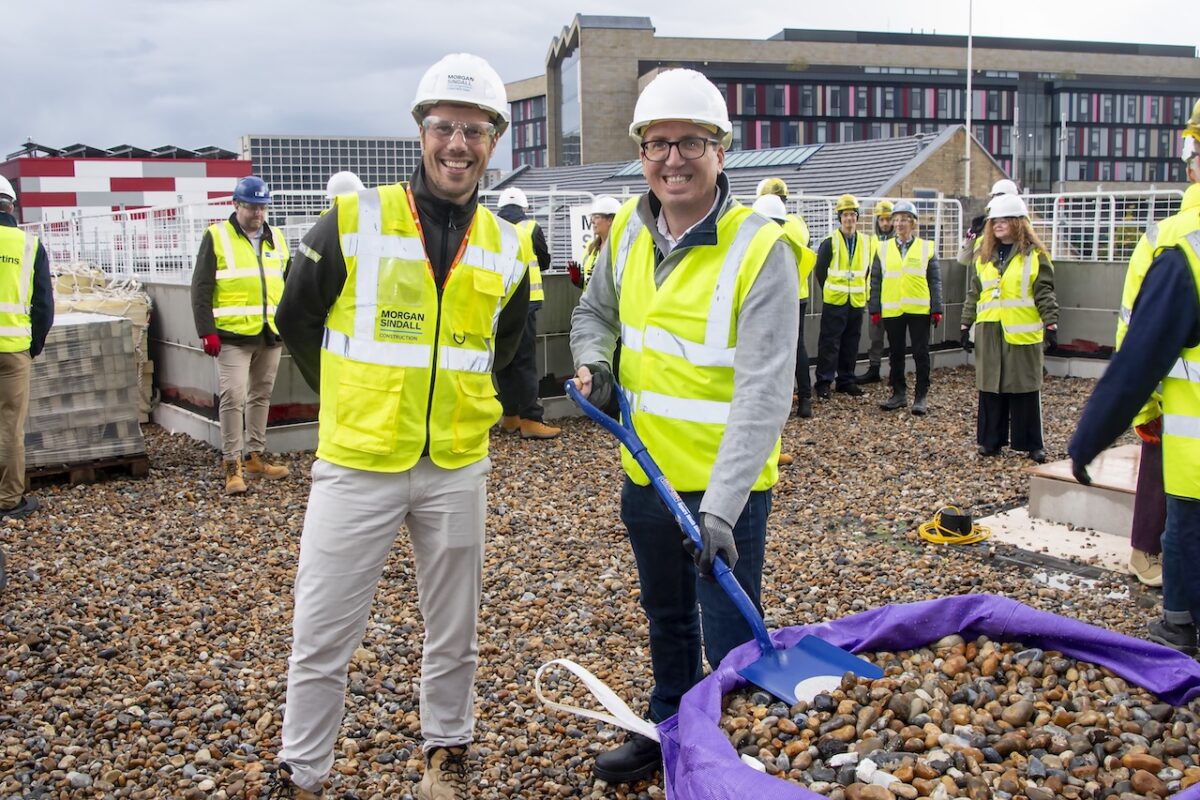 Morgan Sindall Project Manager Dan Marsh stands to the left of Bradford College Vice Principal Chris Malish who is holding a shovel of gravel. They are both on top of the new Junction Mills building with other event attendees in the background. Everyone is wearing high-vis jackets and hard hats.