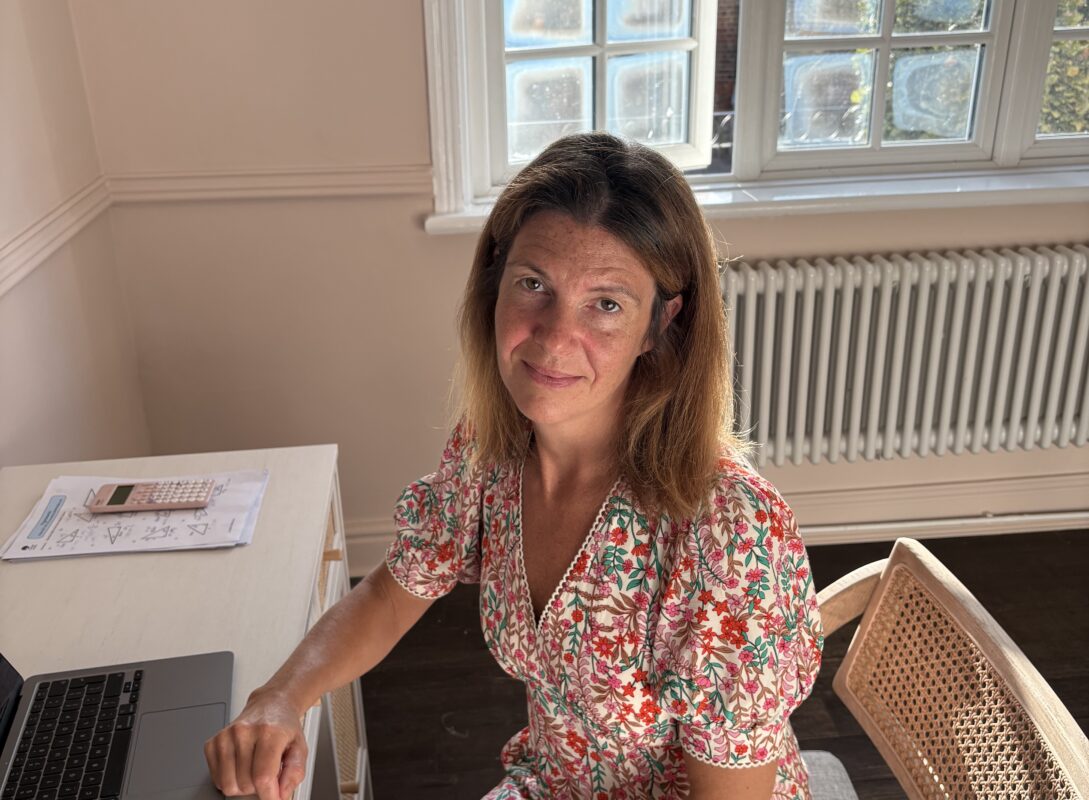 A woman with brown hair sits at a desk smiling at the camera. She is wearing a flowery dress. There is a window behind her letting in lots of natural light.