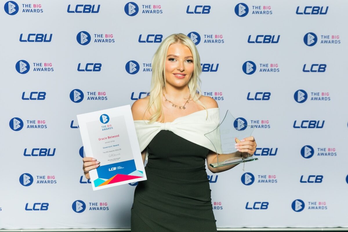 A young, blonde female student stands in front of an LCB banner stand covered in logos, and holds up her glass trophy and certificate while smiling at the camera.