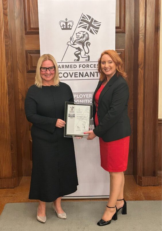 Two women (Jackie Grubb and Pauline Hands) smiling and standing in front of a banner that reads 'Armed Forces Covenant' and jointly holding an ERS award plaque