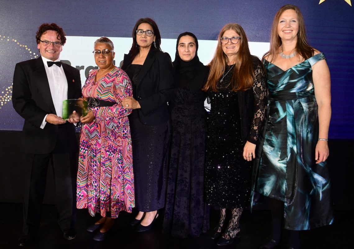 Bradford College staff stand as a group on stage and post with their ‘Benefitting Society’ glass trophy at the 2025 UK & Ireland Green Gown Awards.
