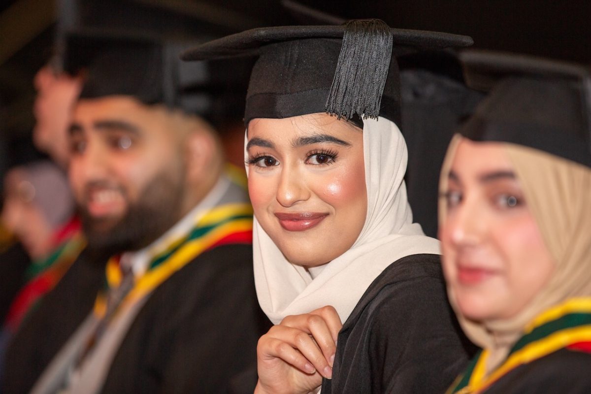 A higher education student smiles at the camera, wearing a tradition graduation gown and a cap over her headscarf. Other graduates are seen to either of her sides.