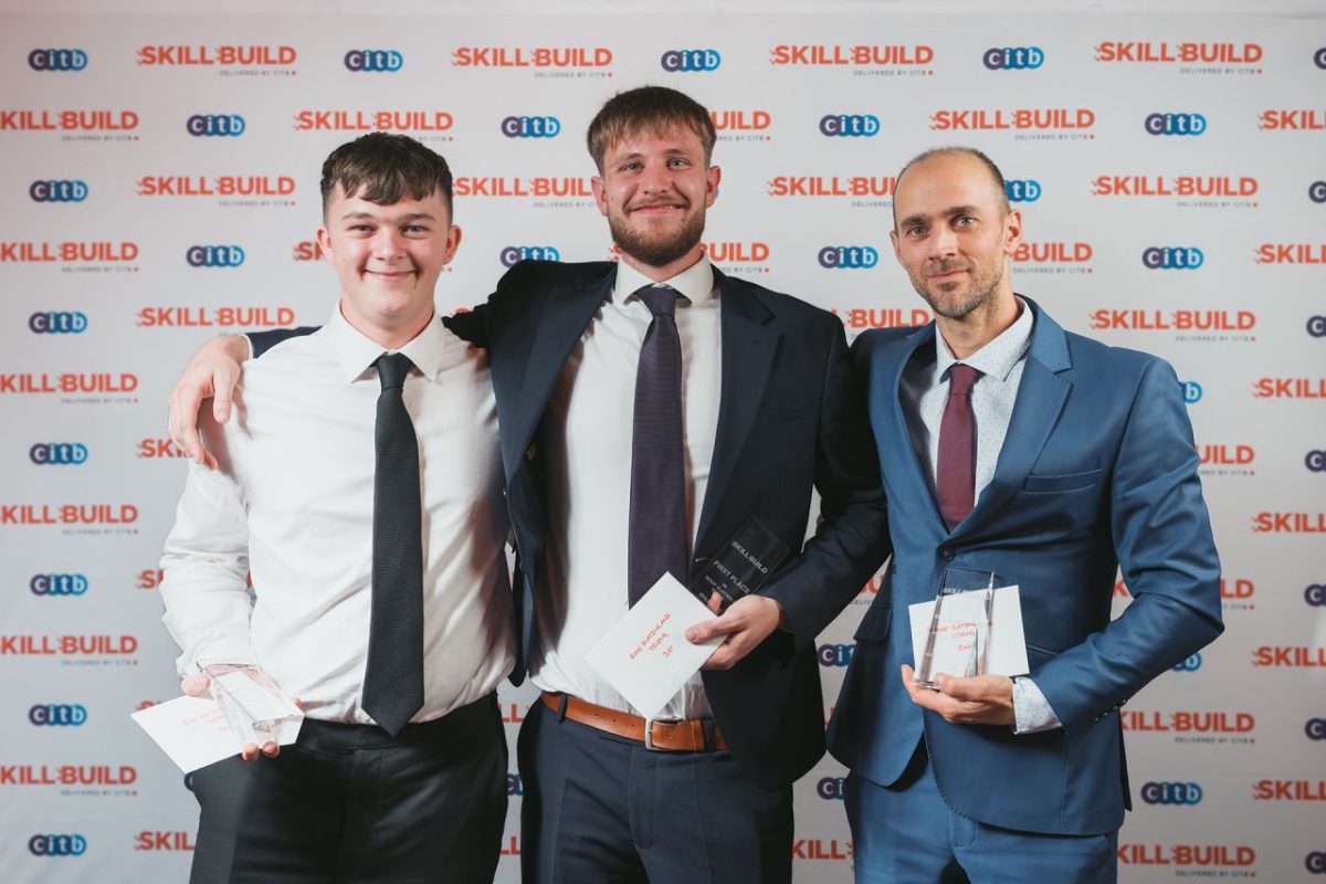 From left to right, Mark Knight (Bronze Medal), Samuel Jinks (Gold Medal), and Levente Szasz (Silver Medal) stand smiling and with arms around each other's shoulders, all holding their SkillBuild 2025 Roof Slating & Tiling trophies. Behind them is a SkillBuild banner stand covered in logos.