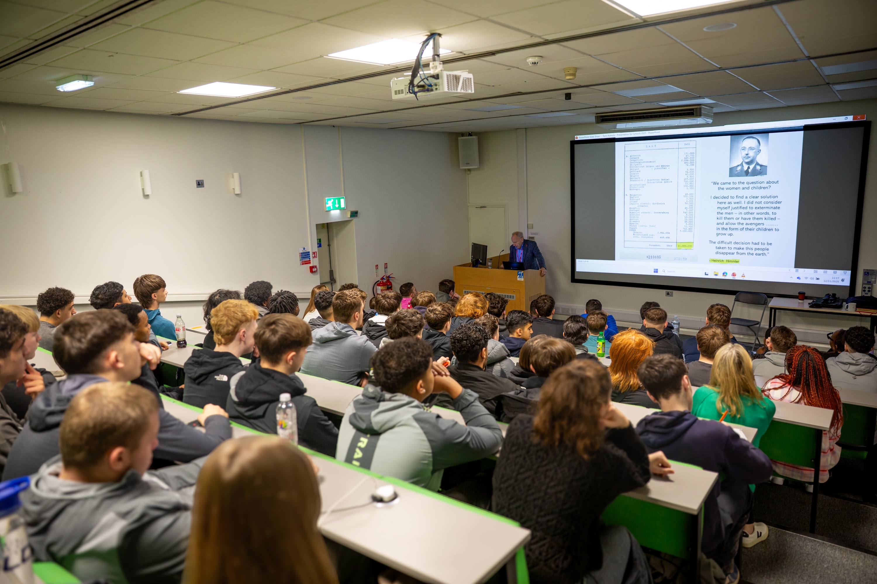 Holocaust survivor, Tomi Komoly, is standing behind a podium at the front of a lecture theatre addressing a full room of LCB English students.