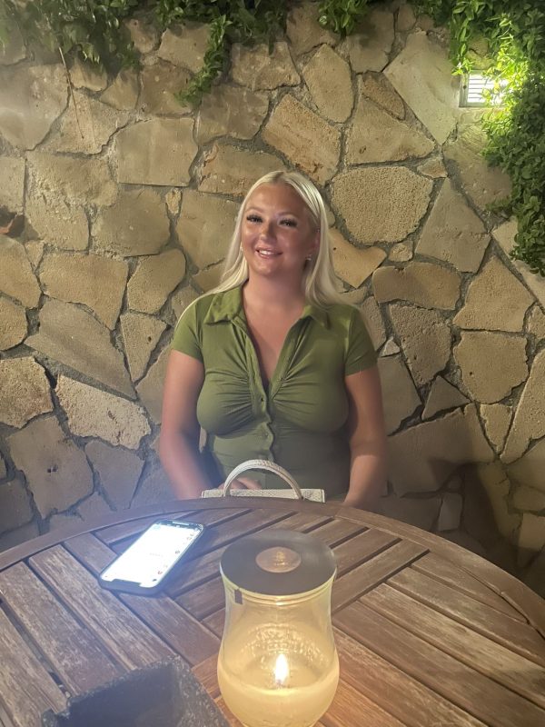 Ella Wilson is in the middle of shot, she has blonde hair, white skin, and is wearing an olive green blouse