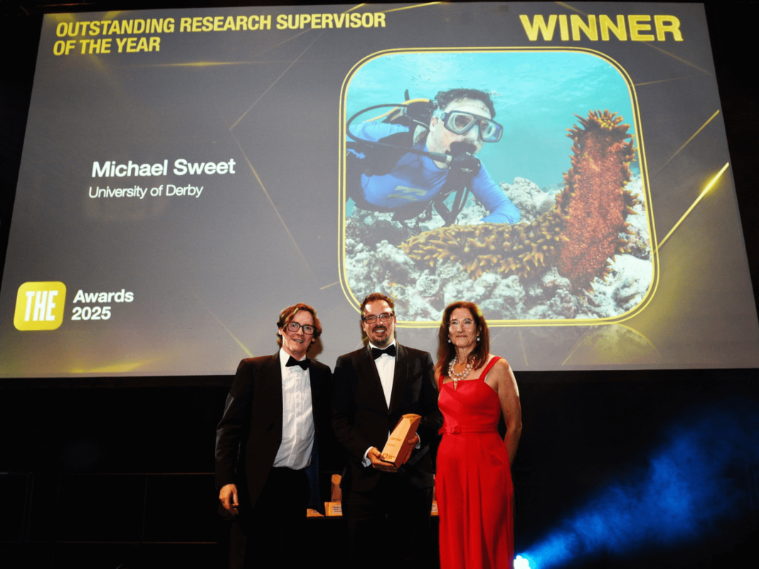 Professor Michael Sweet stands holding an award with two people