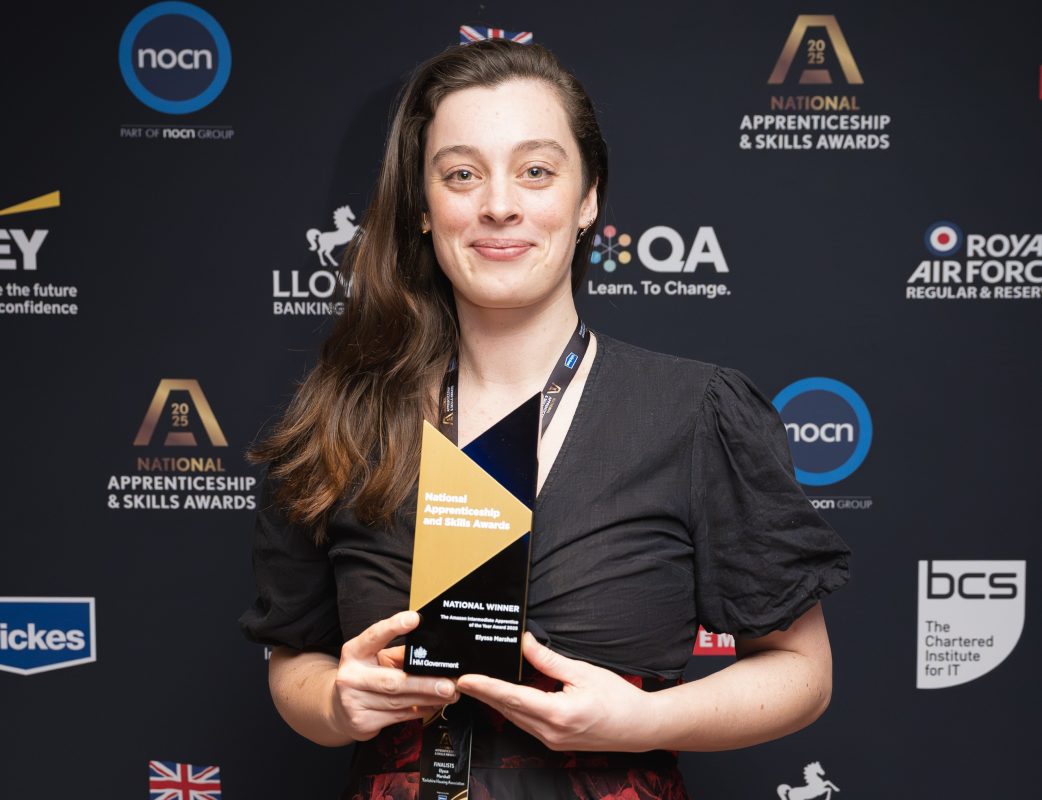 Elyssa Marshall, a Level 2 Property Maintenance Operations apprentice at Yorkshire Housing Association, stands in front of a black backdrop with sponsor logos on it. She is holding her apprenticeship trophy and smiling at the camera. She has a black top on and her long brown hair is swept over one shoulder.