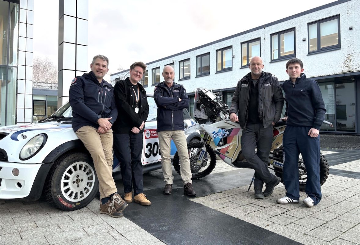 Group photo of 5 people standing in front of a car and motorbike outside a college building