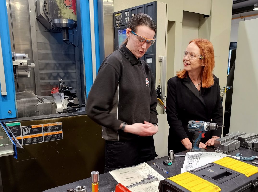 Labour MP for Bradford South, Judith Cummins, stands to the right of a female engineering apprentice at the AESSEAL warehouse. The apprentice is wearing work clothes and eye guards and shows Judith her work station. There are tools on the workbench in front of them.