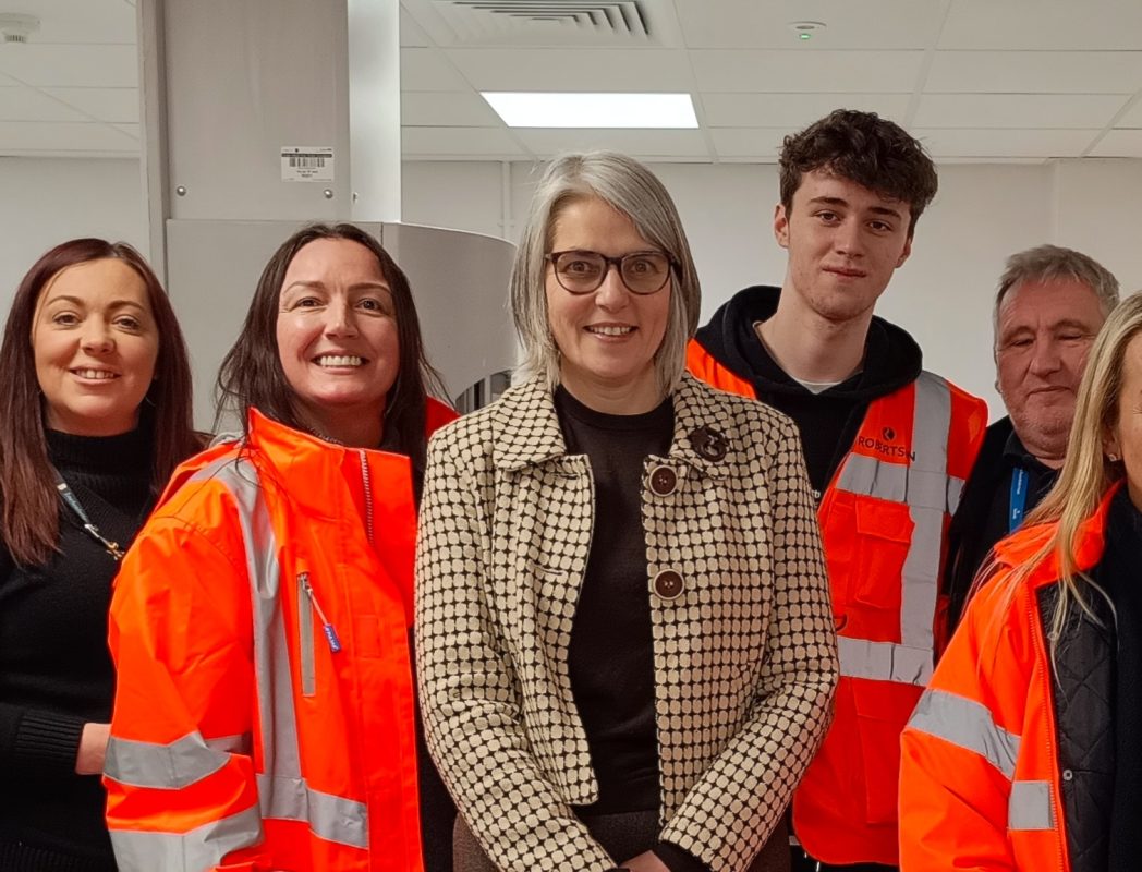 The Labour MP for Shipley, Anna Dixon, stands in the middle of a group of Bradford College T Level students on work placement with Robertson at Airedale General Hospital. Male and female students stand either side of Anna, all wearing bright orange high-vis Robertson work jackets.