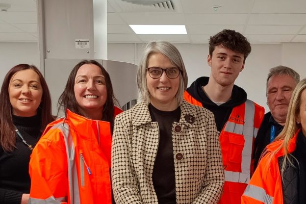 The Labour MP for Shipley, Anna Dixon, stands in the middle of a group of Bradford College T Level students on work placement with Robertson at Airedale General Hospital. Male and female students stand either side of Anna, all wearing bright orange high-vis Robertson work jackets.