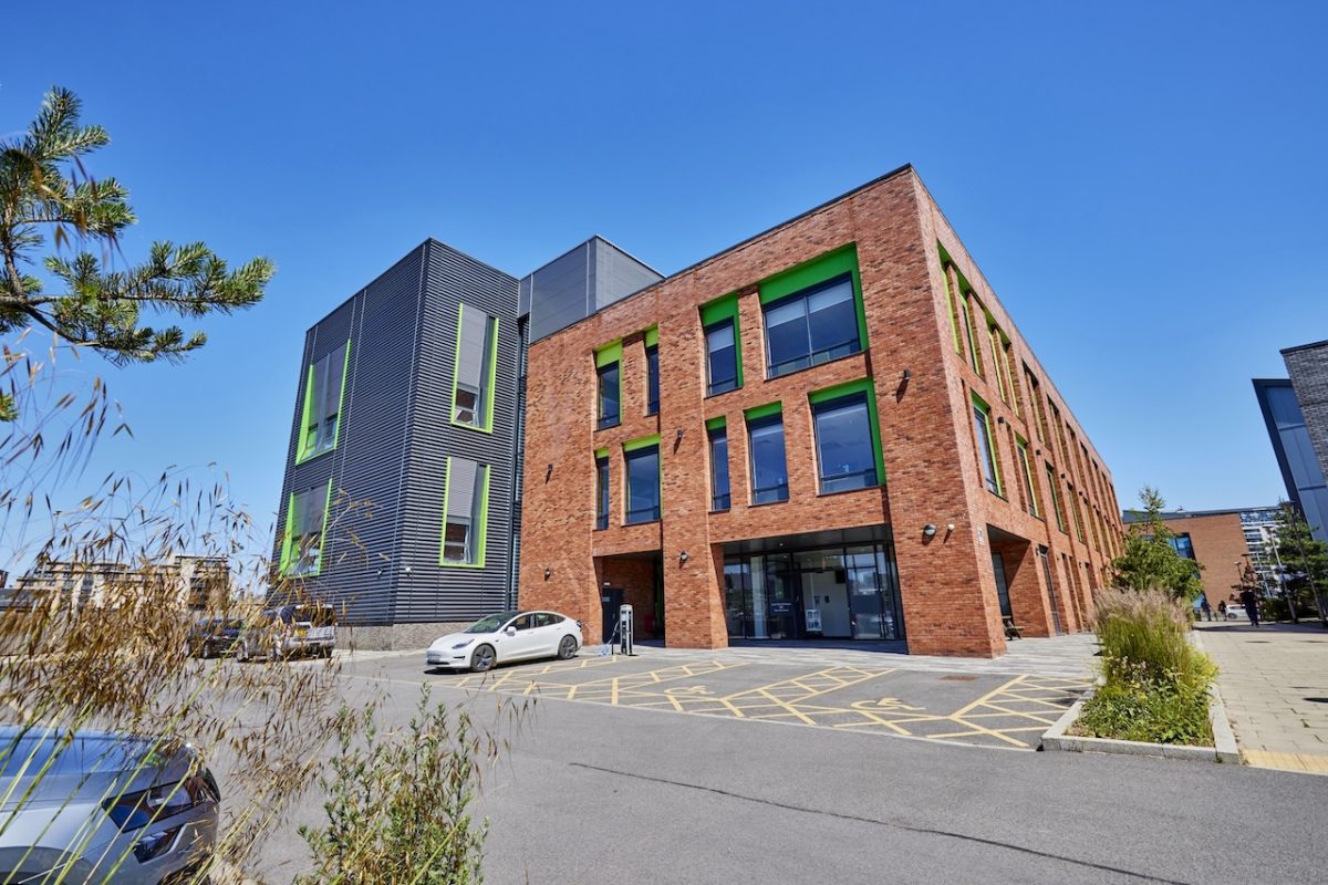 An images of the facade of Leeds College of Building South Bank Campus. It is a three storey red brick building with section clad in grey and green. The sky is clear and blue.