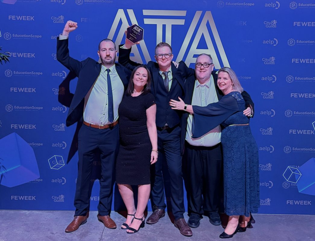 Five members of the Bradford College Apprenticeship team stand in an embrace, celebrating and holding aloft their glass award for 'Apprenticeship Provider of the Year.' They stand in front of a blue backdrop with a large ATA logo on it.