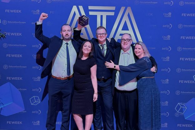 Five members of the Bradford College Apprenticeship team stand in an embrace, celebrating and holding aloft their glass award for 'Apprenticeship Provider of the Year.' They stand in front of a blue backdrop with a large ATA logo on it.