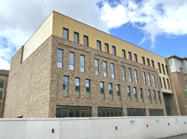 An external shot of the new four-storey building at Bradford College. It is made of brown bricks and a yellow cladding and there are barriers around the bottom as construction work continues.